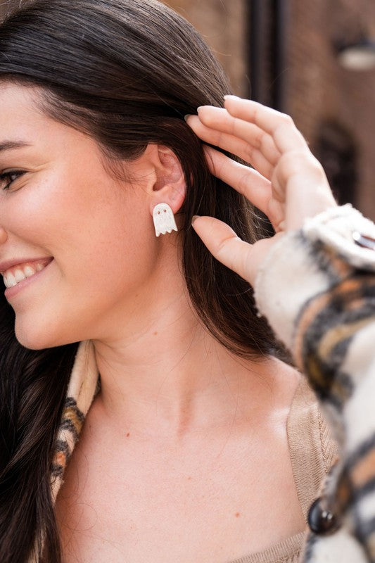 A woman with long dark hair, wearing a beige top and patterned jacket, smiles while showing off her Ghost Studs, small ghost-shaped earrings crafted from hypoallergenic stainless steel.