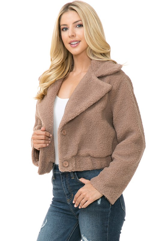 A person is wearing a Women's Faux Fur Jacket in light brown, textured fabric over a white top and blue jeans. They're smiling with their hands in their jacket pockets, exuding an effortlessly chic vibe with the casual solid print of the outfit.