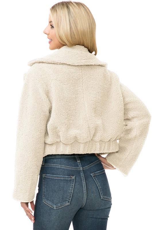 A person is wearing a Women's Faux Fur Jacket in light brown, textured fabric over a white top and blue jeans. They're smiling with their hands in their jacket pockets, exuding an effortlessly chic vibe with the casual solid print of the outfit.