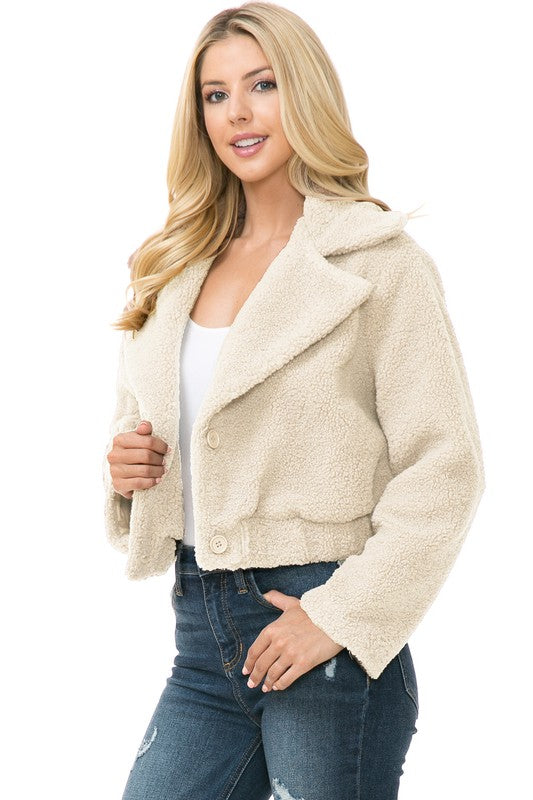 A person is wearing a Women's Faux Fur Jacket in light brown, textured fabric over a white top and blue jeans. They're smiling with their hands in their jacket pockets, exuding an effortlessly chic vibe with the casual solid print of the outfit.