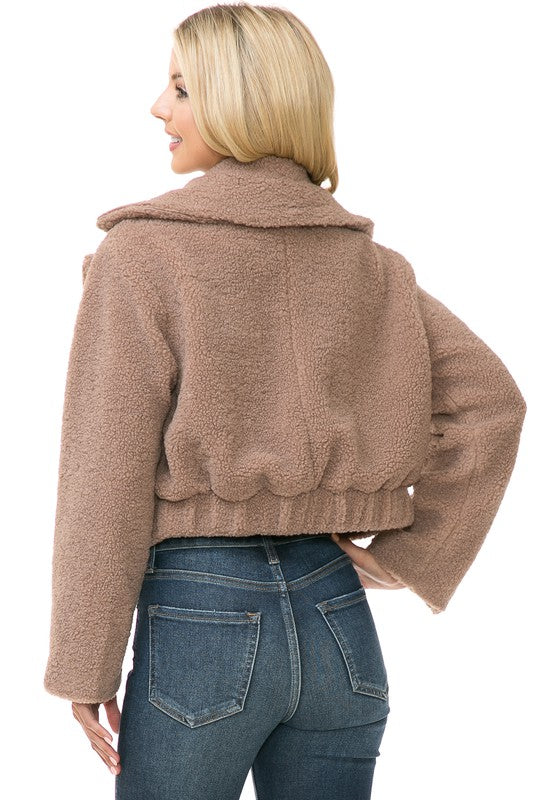 A person is wearing a Women's Faux Fur Jacket in light brown, textured fabric over a white top and blue jeans. They're smiling with their hands in their jacket pockets, exuding an effortlessly chic vibe with the casual solid print of the outfit.
