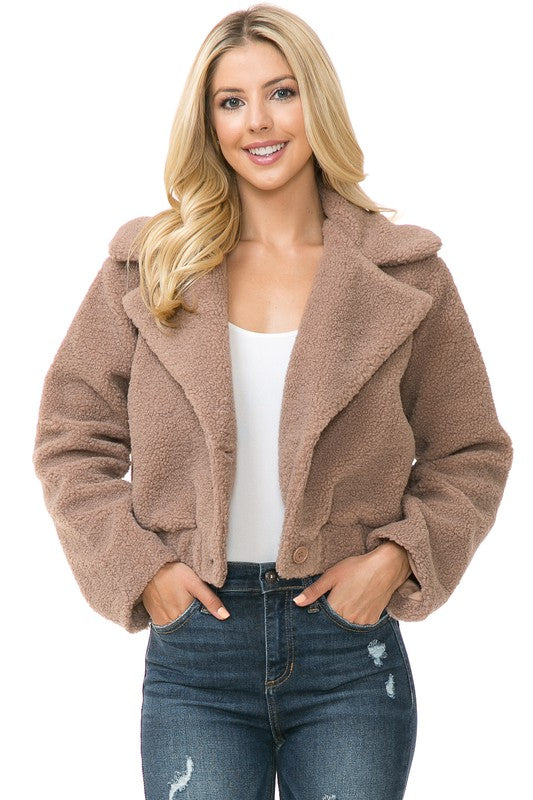 A person is wearing a Women's Faux Fur Jacket in light brown, textured fabric over a white top and blue jeans. They're smiling with their hands in their jacket pockets, exuding an effortlessly chic vibe with the casual solid print of the outfit.