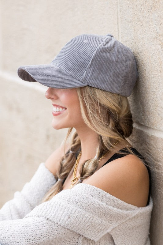 Sentence with the replaced product name: Woman smiling and leaning against a wall while holding the brim of her Corduroy Boyfriend Ball Cap.