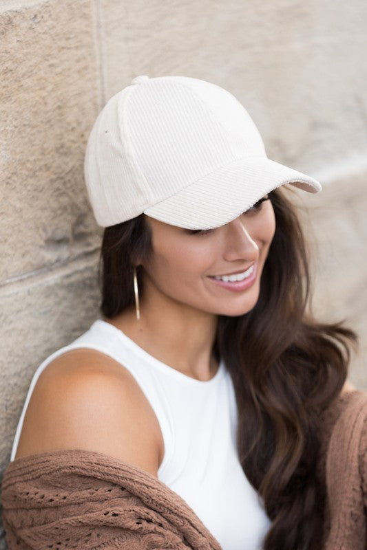Sentence with the replaced product name: Woman smiling and leaning against a wall while holding the brim of her Corduroy Boyfriend Ball Cap.