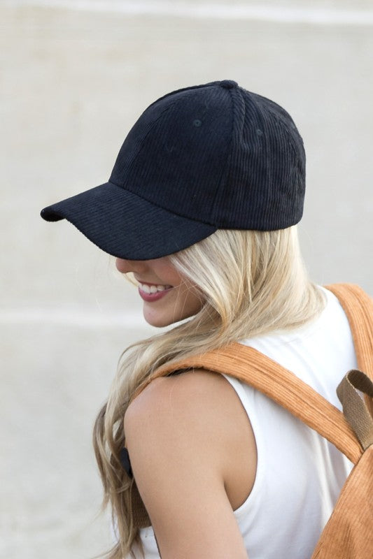 Sentence with the replaced product name: Woman smiling and leaning against a wall while holding the brim of her Corduroy Boyfriend Ball Cap.