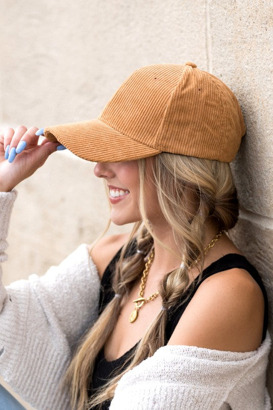 Sentence with the replaced product name: Woman smiling and leaning against a wall while holding the brim of her Corduroy Boyfriend Ball Cap.