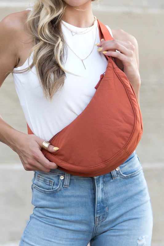 A woman holding an Everyday Sling bag with an adjustable strap length across her body while wearing a white tank top and blue jeans. Only her torso and the bag are visible.