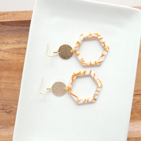 Close-up of a person wearing the Lennox Earrings - Pumpkin Spice, featuring a brown and white hexagonal design with a round gold disc, paired with a brown sweater.