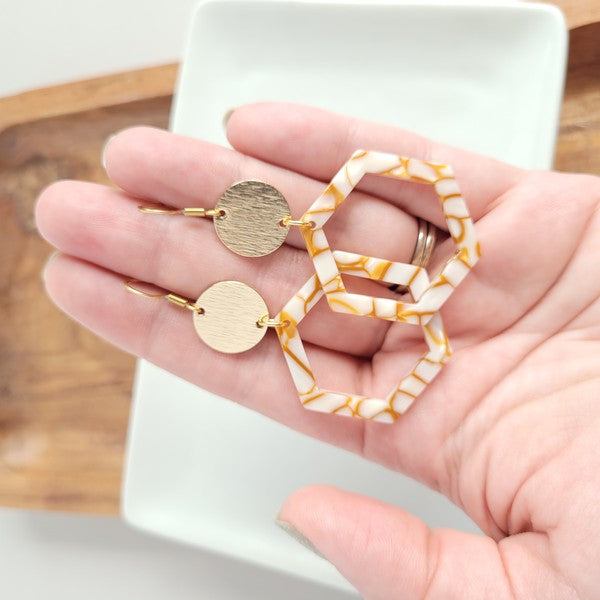 Close-up of a person wearing the Lennox Earrings - Pumpkin Spice, featuring a brown and white hexagonal design with a round gold disc, paired with a brown sweater.