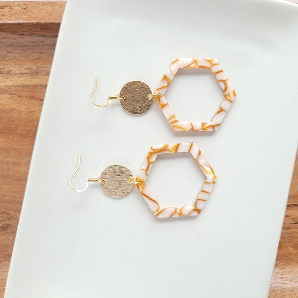 Close-up of a person wearing the Lennox Earrings - Pumpkin Spice, featuring a brown and white hexagonal design with a round gold disc, paired with a brown sweater.