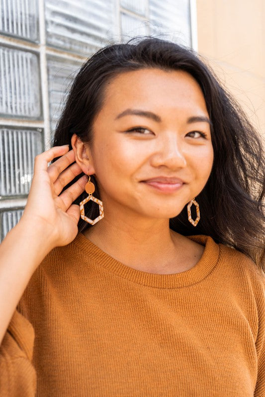 Close-up of a person wearing the Lennox Earrings - Pumpkin Spice, featuring a brown and white hexagonal design with a round gold disc, paired with a brown sweater.
