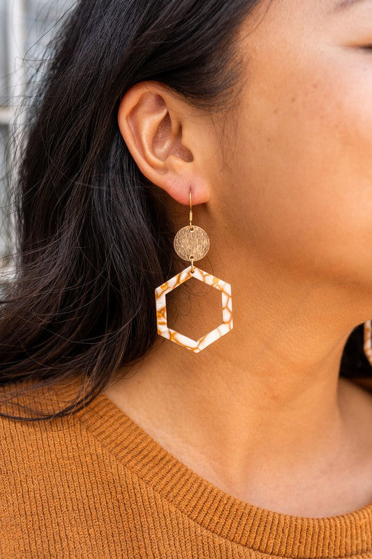 Close-up of a person wearing the Lennox Earrings - Pumpkin Spice, featuring a brown and white hexagonal design with a round gold disc, paired with a brown sweater.