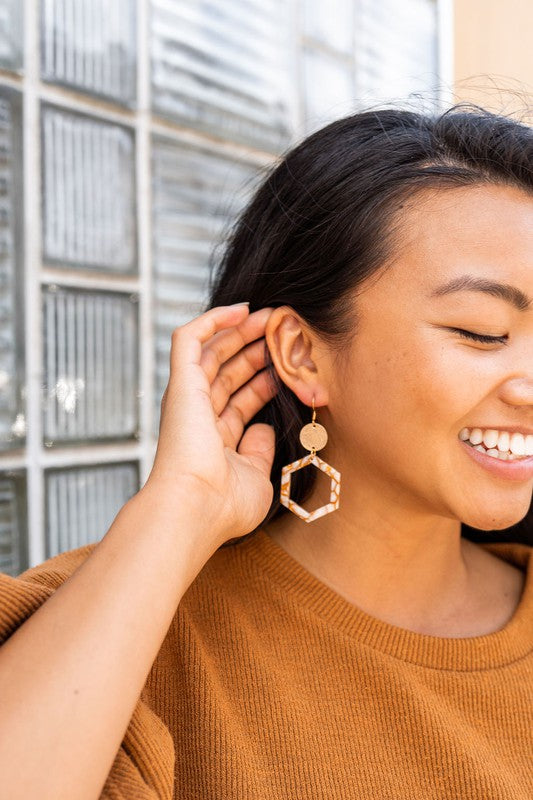 Close-up of a person wearing the Lennox Earrings - Pumpkin Spice, featuring a brown and white hexagonal design with a round gold disc, paired with a brown sweater.