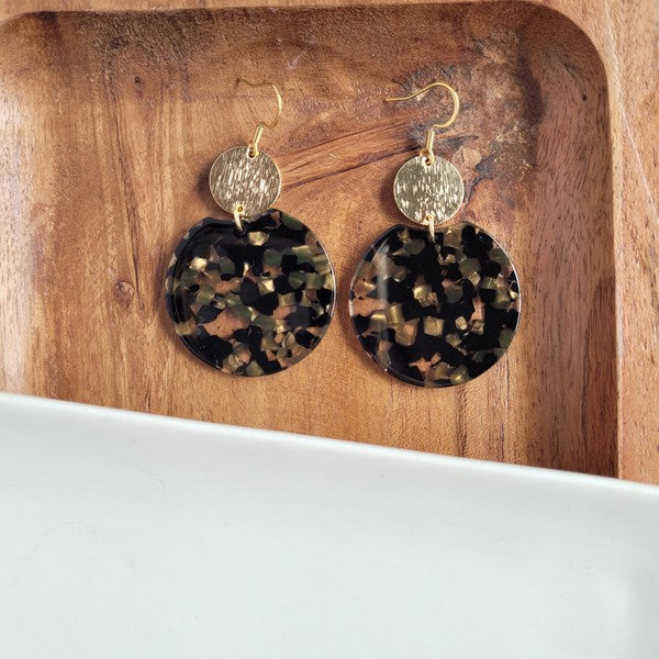 A pair of Zoey Earrings - Black & Bronze, adorned with 18K gold-plated hooks and featuring a large black, beige, and clear speckled disc with a smaller textured bronze disc above it, elegantly displayed on a white tray.