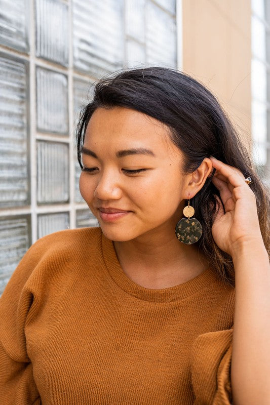 A pair of Zoey Earrings - Black & Bronze, adorned with 18K gold-plated hooks and featuring a large black, beige, and clear speckled disc with a smaller textured bronze disc above it, elegantly displayed on a white tray.