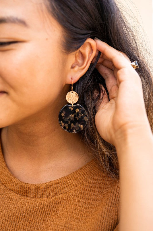 A pair of Zoey Earrings - Black & Bronze, adorned with 18K gold-plated hooks and featuring a large black, beige, and clear speckled disc with a smaller textured bronze disc above it, elegantly displayed on a white tray.