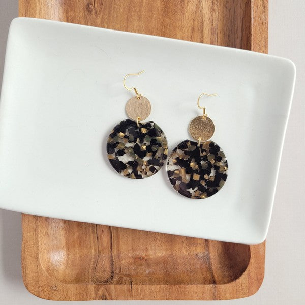 A pair of Zoey Earrings - Black & Bronze, adorned with 18K gold-plated hooks and featuring a large black, beige, and clear speckled disc with a smaller textured bronze disc above it, elegantly displayed on a white tray.