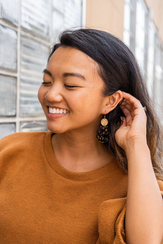 A pair of Zoey Earrings - Black & Bronze, adorned with 18K gold-plated hooks and featuring a large black, beige, and clear speckled disc with a smaller textured bronze disc above it, elegantly displayed on a white tray.