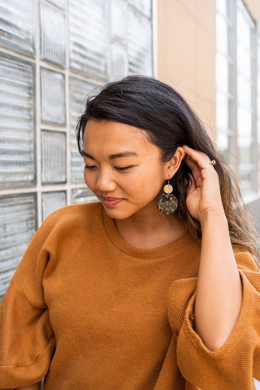 A pair of Zoey Earrings - Black & Bronze, adorned with 18K gold-plated hooks and featuring a large black, beige, and clear speckled disc with a smaller textured bronze disc above it, elegantly displayed on a white tray.