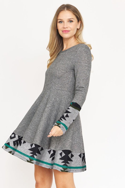 A woman wearing the Layered Fit And Flare Dress, featuring gray long sleeves and black and green geometric patterns, stands against a plain white background.