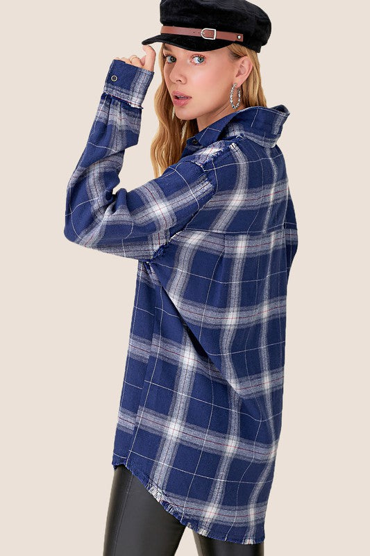 A person wearing the Celine Shirt smiles at the camera, their long, wavy hair cascading down as they stand against a beige background. The blue checkered plaid flannel shirt features a button-front closure, which they highlight with one hand on their hip. Complementing the look is a black cap that completes their stylish ensemble.