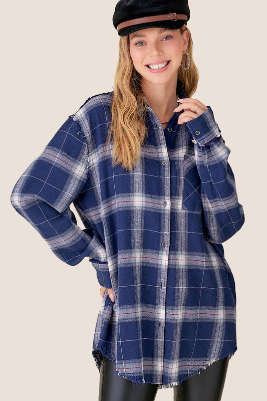 A woman wearing the Celine Shirt, a long sleeve checkered plaid flannel with a button-front closure, along with a black cap and black pants, stands smiling with one hand on her hip against a plain background.