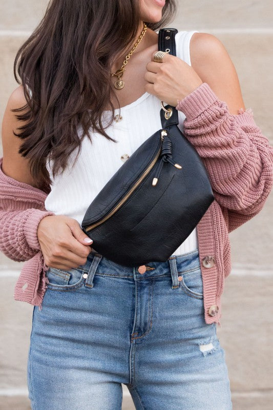 A person wearing light blue jeans and a white tank top is holding a Luxe Convertible Sling Belt Bum Bag with a zippered pocket.