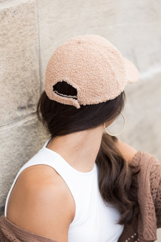 A smiling woman in a white top and Boucle Sherpa Teddy Bear knit ball cap against a stone wall.