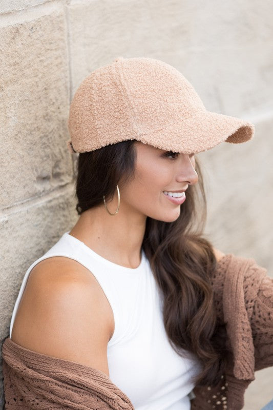A smiling woman in a white top and Boucle Sherpa Teddy Bear knit ball cap against a stone wall.