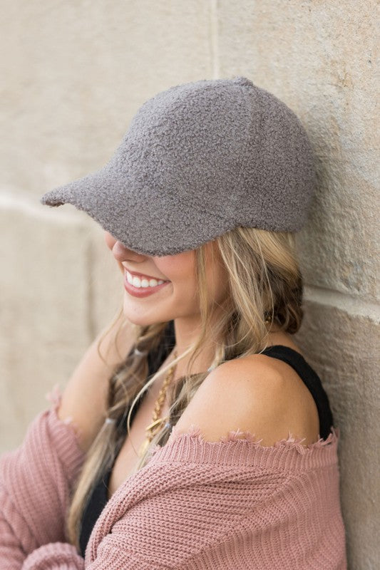A smiling woman in a white top and Boucle Sherpa Teddy Bear knit ball cap against a stone wall.