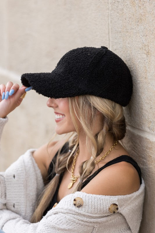 A smiling woman in a white top and Boucle Sherpa Teddy Bear knit ball cap against a stone wall.