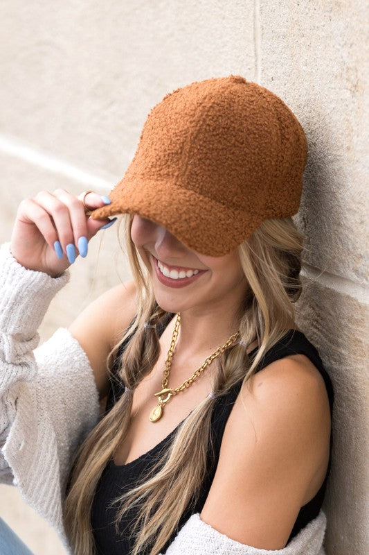 A smiling woman in a white top and Boucle Sherpa Teddy Bear knit ball cap against a stone wall.