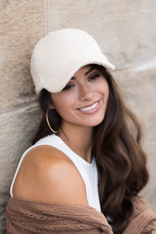 A smiling woman in a white top and Boucle Sherpa Teddy Bear knit ball cap against a stone wall.