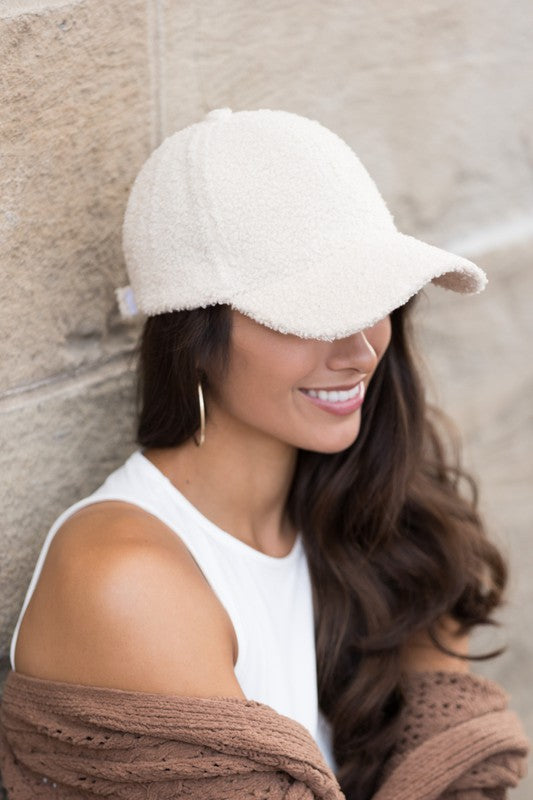 A smiling woman in a white top and Boucle Sherpa Teddy Bear knit ball cap against a stone wall.