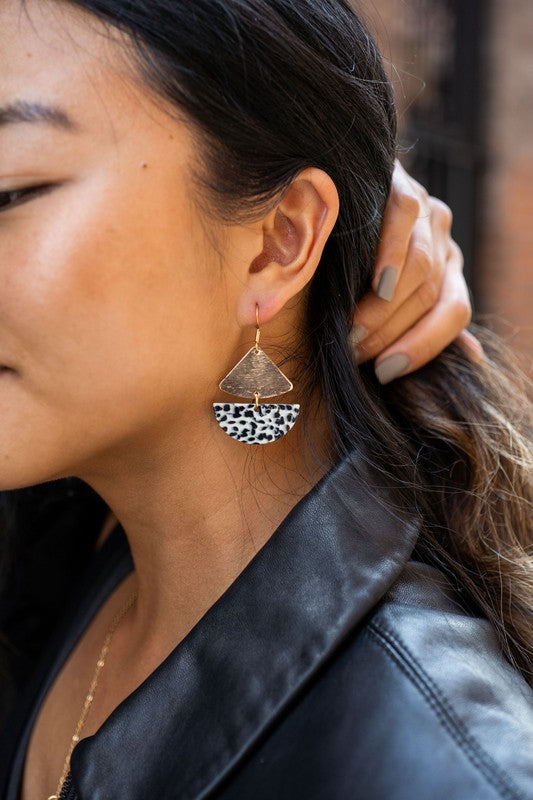 Close-up of a person wearing the Ava Black Dot earrings, featuring gold-plated hooks and a distinctive leopard print. Their dark hair partially obscures their ear while their hand, adorned with neutral-colored nail polish, rests behind their neck.