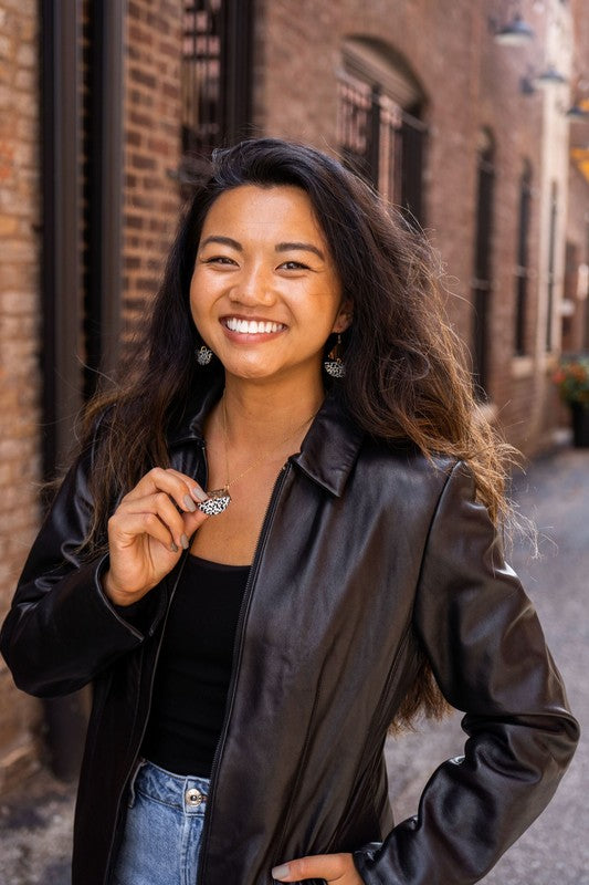 Close-up of a person wearing the Ava Black Dot earrings, featuring gold-plated hooks and a distinctive leopard print. Their dark hair partially obscures their ear while their hand, adorned with neutral-colored nail polish, rests behind their neck.