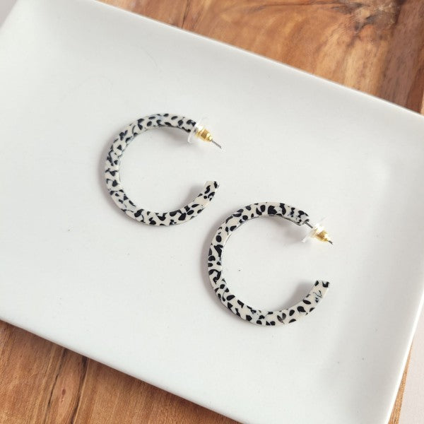 The Camy Hoops - Black Dot are medium-sized hoop earrings featuring a black and white spotted pattern, crafted from lightweight acrylics, elegantly displayed on a white rectangular dish placed on a wooden surface.