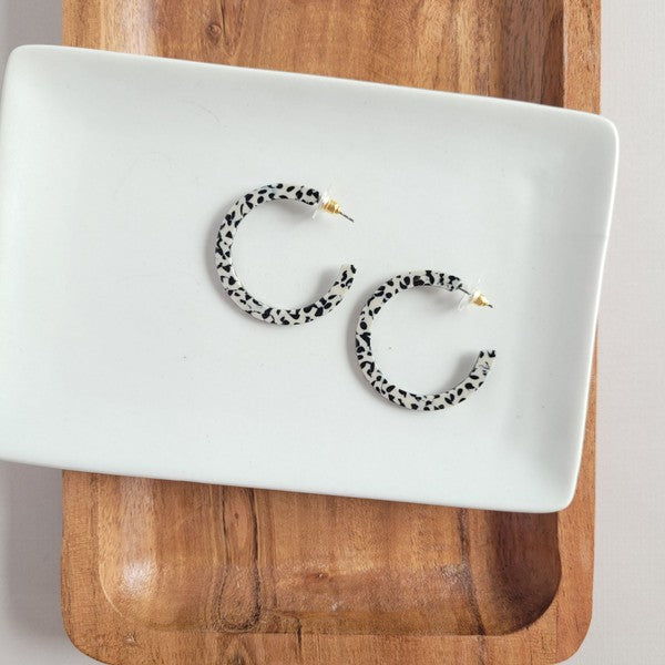 The Camy Hoops - Black Dot are medium-sized hoop earrings featuring a black and white spotted pattern, crafted from lightweight acrylics, elegantly displayed on a white rectangular dish placed on a wooden surface.