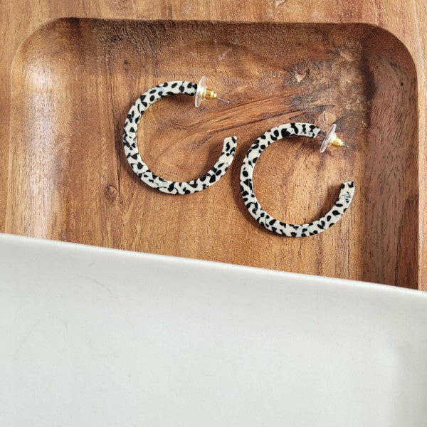 The Camy Hoops - Black Dot are medium-sized hoop earrings featuring a black and white spotted pattern, crafted from lightweight acrylics, elegantly displayed on a white rectangular dish placed on a wooden surface.