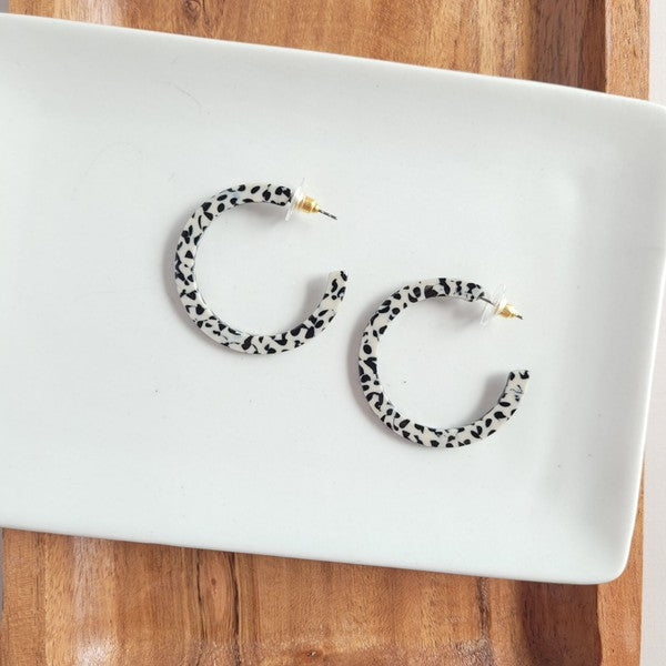 The Camy Hoops - Black Dot are medium-sized hoop earrings featuring a black and white spotted pattern, crafted from lightweight acrylics, elegantly displayed on a white rectangular dish placed on a wooden surface.