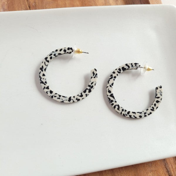 The Camy Hoops - Black Dot are medium-sized hoop earrings featuring a black and white spotted pattern, crafted from lightweight acrylics, elegantly displayed on a white rectangular dish placed on a wooden surface.