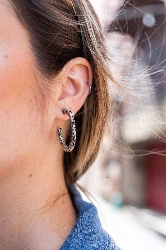 The Camy Hoops - Black Dot are medium-sized hoop earrings featuring a black and white spotted pattern, crafted from lightweight acrylics, elegantly displayed on a white rectangular dish placed on a wooden surface.