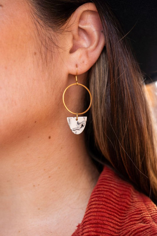 Close-up of a woman's ear adorned with the Iris Earrings - Marble, featuring dainty dangle earrings with an 18K gold-plated hoop and a white marble-patterned pendant. She has long brown hair and is wearing a red textured top.