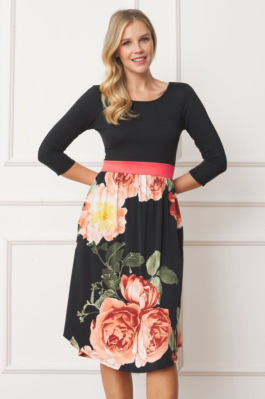 A woman in a Curved Hem Band Midi Dress, featuring a navy blue top, yellow waistband, and floral skirt, stands against a white wall, smiling.