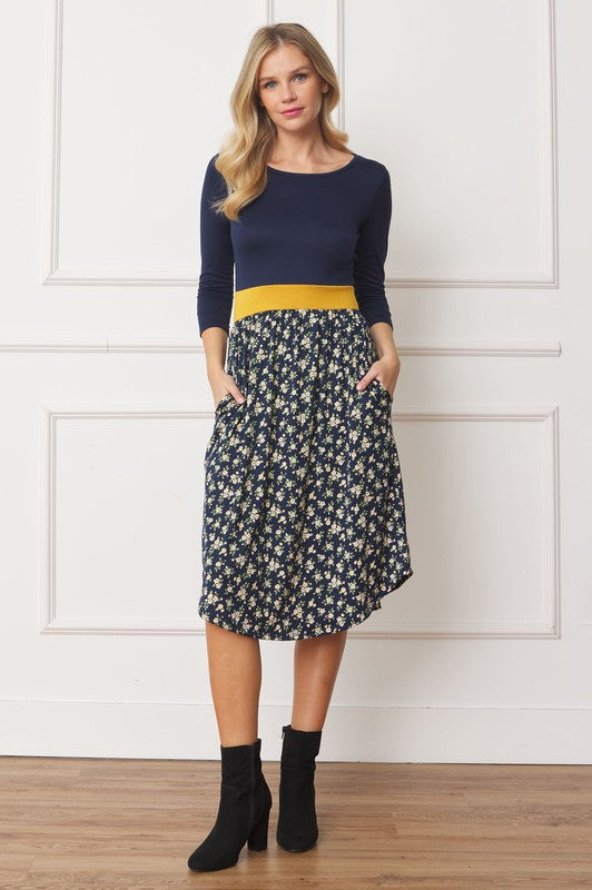 A woman in a Curved Hem Band Midi Dress, featuring a navy blue top, yellow waistband, and floral skirt, stands against a white wall, smiling.