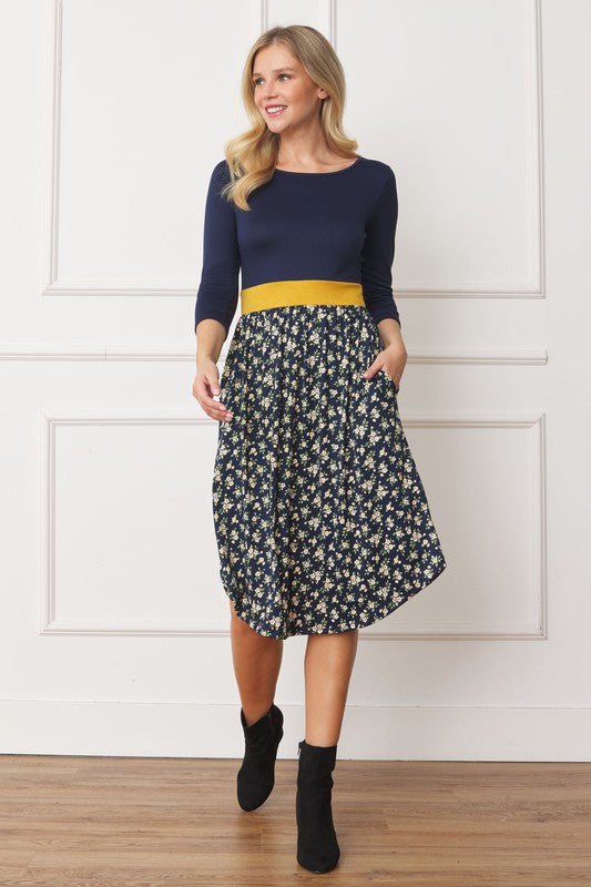A woman in a Curved Hem Band Midi Dress, featuring a navy blue top, yellow waistband, and floral skirt, stands against a white wall, smiling.