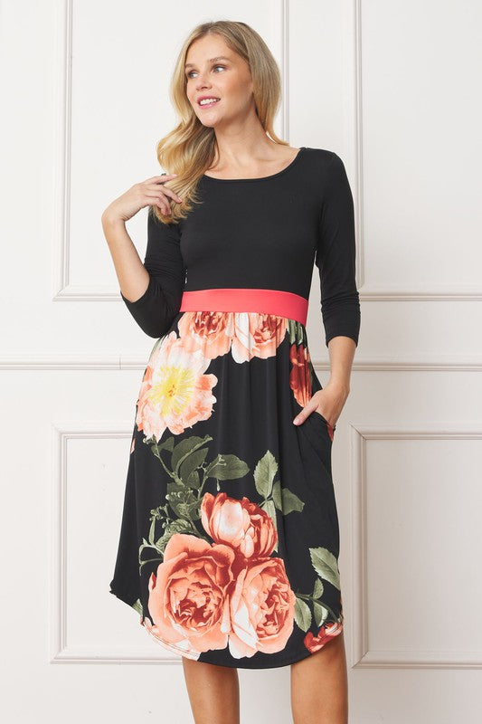 A woman in a Curved Hem Band Midi Dress, featuring a navy blue top, yellow waistband, and floral skirt, stands against a white wall, smiling.