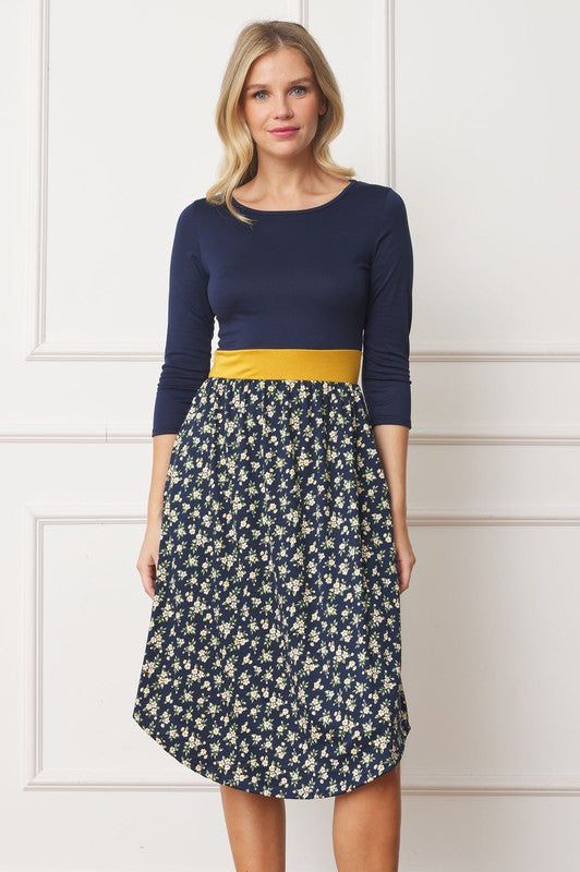 A woman in a Curved Hem Band Midi Dress, featuring a navy blue top, yellow waistband, and floral skirt, stands against a white wall, smiling.