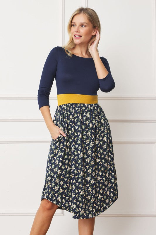 A woman in a Curved Hem Band Midi Dress, featuring a navy blue top, yellow waistband, and floral skirt, stands against a white wall, smiling.