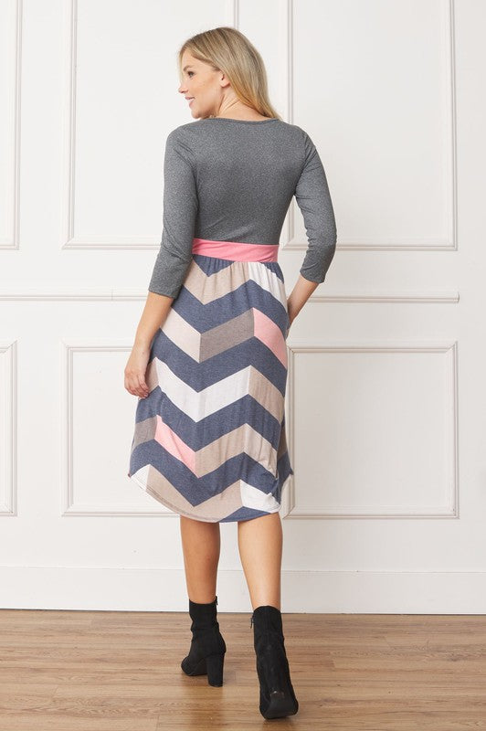 A woman in a Curved Hem Band Midi Dress, featuring a navy blue top, yellow waistband, and floral skirt, stands against a white wall, smiling.